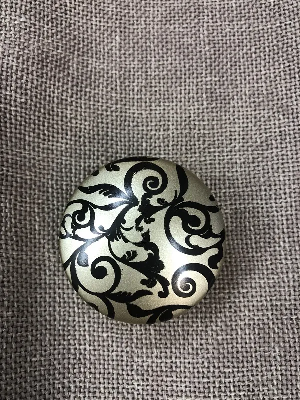 Круглый декоративный подхват с черно-серебристым узором лежит на серой льняной ткани. Виден роскошный орнамент и матовая текстура. Подходит для оконных аксессуаров в классическом стиле.