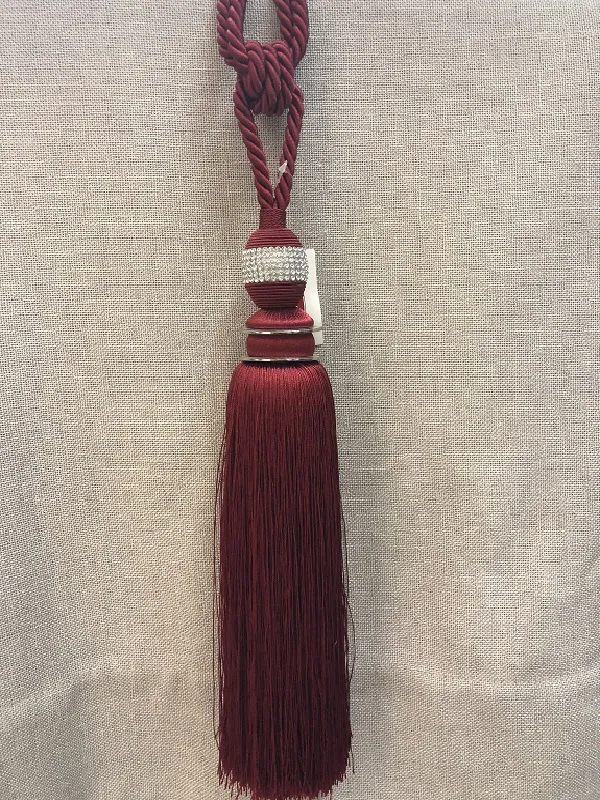Роскошный бордовый подхват с кистью и бусинами висит на фоне ткани. Декоративный элемент для штор, создающий атмосферу утончённости и роскоши в интерьере.