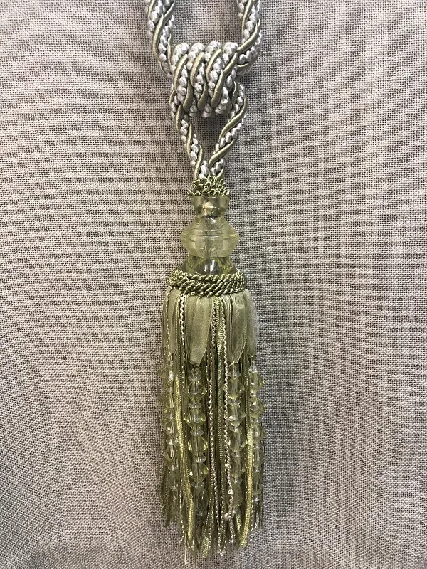 Золотистая кисть с бусинами висит на тканевом фоне. Свет мягко проникает сквозь ткань, подчёркивая текстуру и блеск украшения. Уютная атмосфера, идеально впишется в классический или современный интерьер.