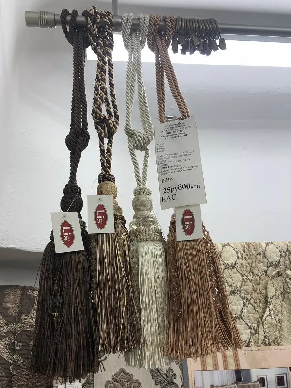 На металлическом карнизе висят четыре кисти из плетёной ткани в тёплых оттенках — коричневом, бежевом и кремовом. Они мягко колышутся, создавая уютную атмосферу. На заднем плане — ткань с узором, подчёркивающая роскошь аксессуаров.