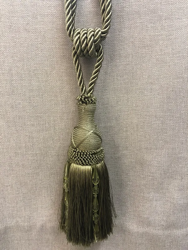 Золотистая кисть для штор, висящая на фоне тёмно-зелёной ткани. Интерьер в классическом стиле с мягким естественным освещением. Декоративный акцент, добавляющий утончённости и роскоши оконному оформлению.