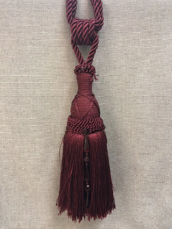 Тёмно-бордовая кисть для штор лежит на тканевом фоне, мягкий свет подчёркивает фактуру шнура и блеск подвески, создаёт атмосферу утончённого уюта и роскоши.