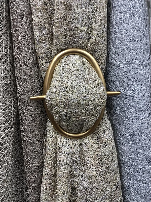 Золотистый подхват с круглым кольцом аккуратно держит складки тонкой серой ткани. Интерьер в спокойных тонах, мягкий рассеянный свет, уютная атмосфера.