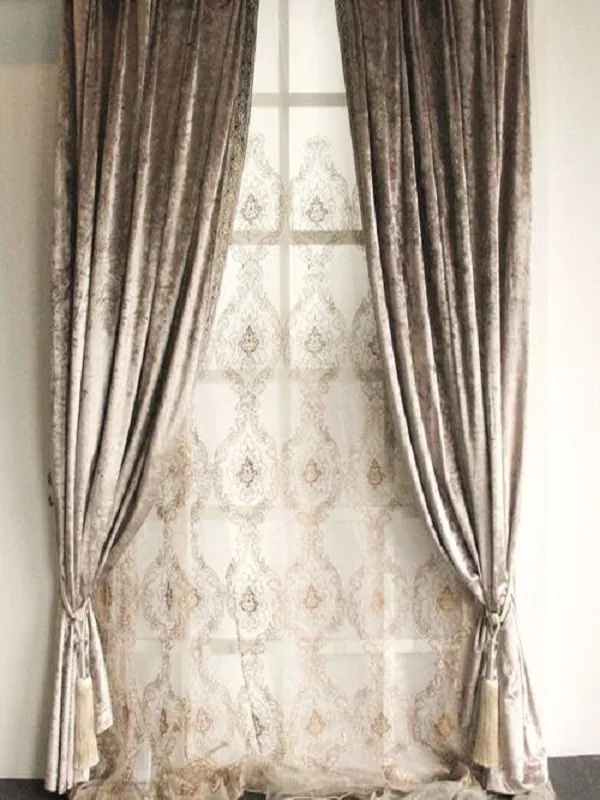 Velvet curtains in dusty taupe with gold lace overlay, elegantly tied back to reveal patterned sheers, soft natural light filtering through in a classic living room setting.