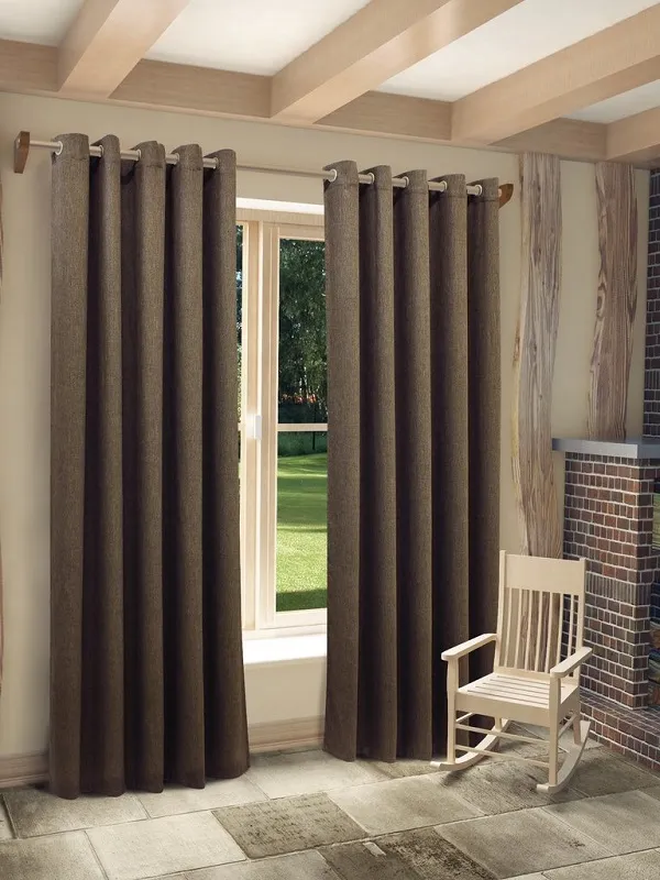 Тяжёлые бархатные шторы в тёмно-коричневом оттенке висят на люверсах, мягко скрывают окно с видом на зелёный сад. Интерьер в стиле лофт с деревянными балками, кирпичной печью и белой кресло-качалкой. Теплое естественное освещение, уютная атмосфера.