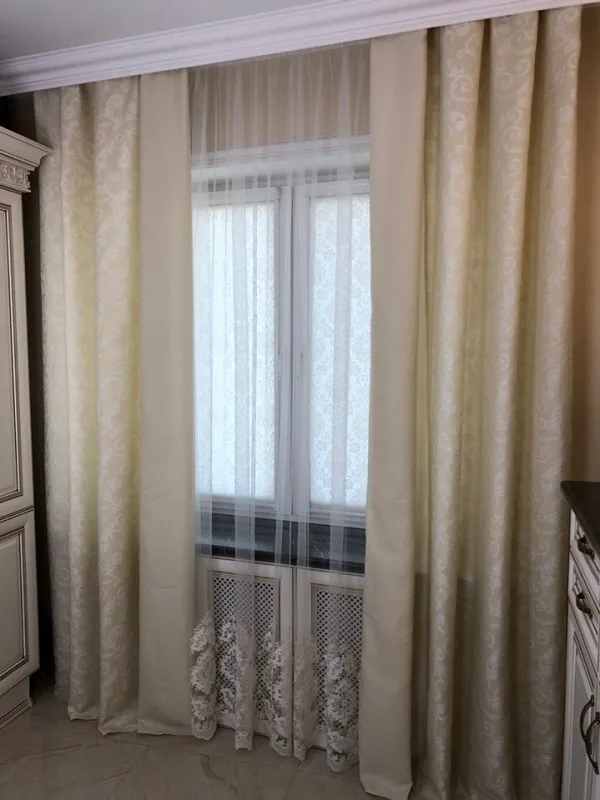 Классический интерьер с пышными шторами в кремовом оттенке и тонким узором, мягкий дневной свет проникает через тюль, создаёт уютную атмосферу, рядом видны элементы мебели в стиле прованс.