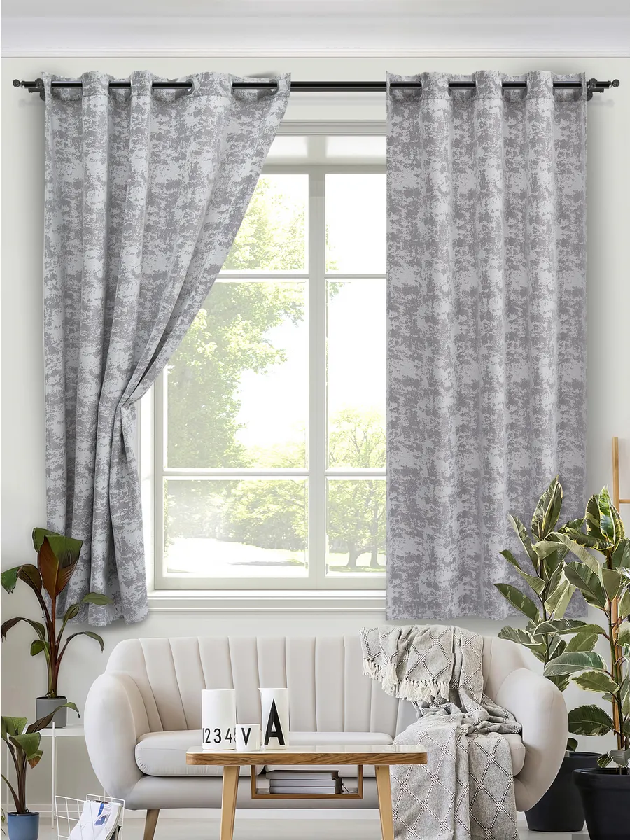 Серые шторы на люверсах в светлой гостиной, мягко падают по обе стороны от окна, за окном зелень деревьев, уютный диван с пледом и зелёные растения по бокам.