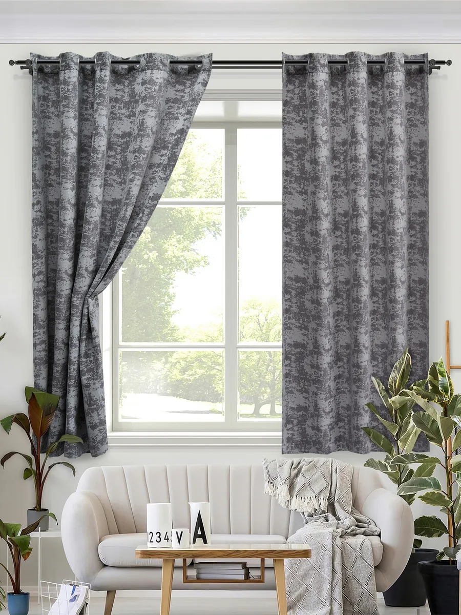 Серые шторы на люверсах висят у окна, мягко складываясь. Свет проходит сквозь ткань, создавая рассеянный уют. В комнате — белый диван, деревянный стол и зелёные растения. Атмосфера спокойной элегантности.