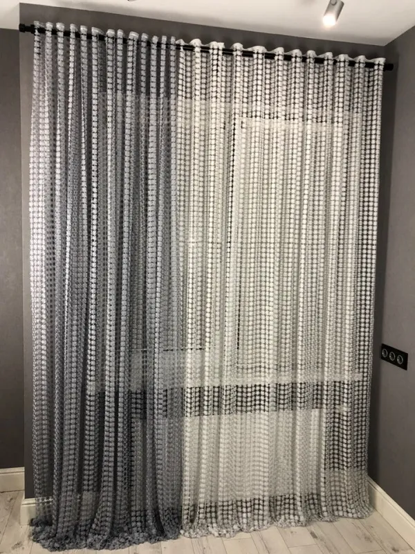 Modern geometric mesh curtain with grey and white grid pattern, softly diffusing daylight in a minimalist urban loft interior with light wood floors and grey walls.