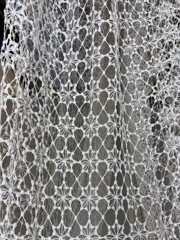 Воздушная сетчатая штора с геометрическим узором висит на окне, пропуская рассеянный свет. Интерьер выглядит уютно и современно. Ткань колышется, создавая игру света и тени.