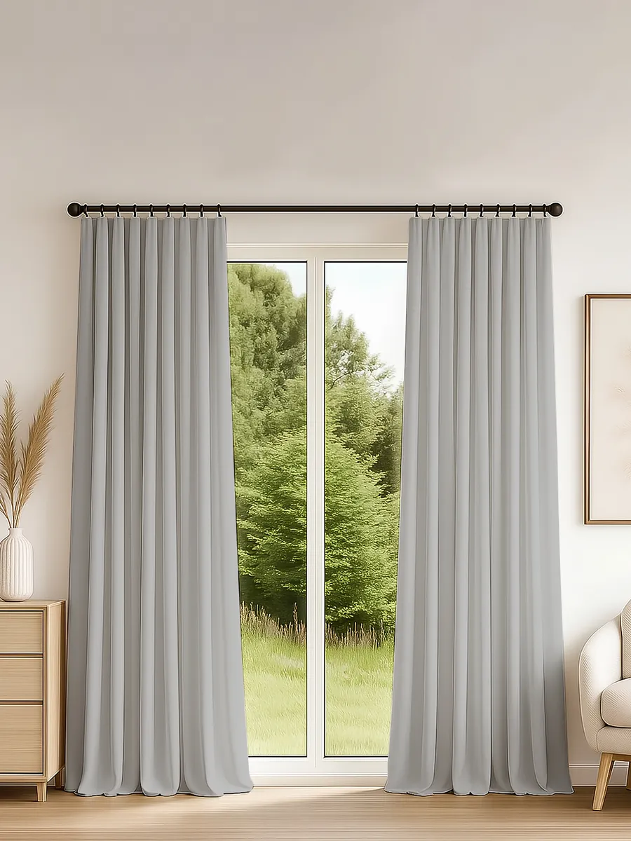 Серые шторы на ленте висят у большого окна, за которым виднеется зелёный лес. Интерьер выполнен в светлых тонах с деревянной мебелью и нейтральной стеной. Ткань мягко падает складками, создавая ощущение уюта и спокойствия.