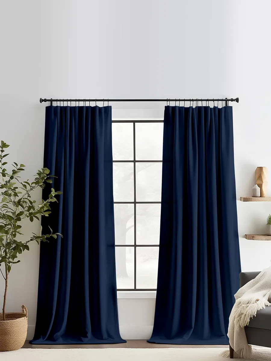 Тяжёлые синие шторы на ленте висят на чёрном карнизе, мягко приглушая свет из окна с чёрными рамами. Интерьер в белых тонах с зелёным растением в плетёном горшке и серым диваном с белым пледом.