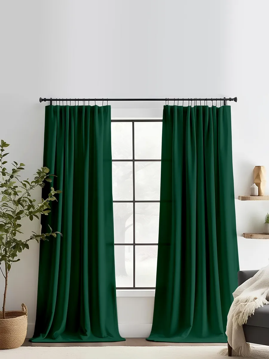 Тяжёлые зелёные шторы на ленте висят на чёрном карнизе, мягко складываясь у окна с чёрными рамами. В комнате — светлые стены, растение в плетёном горшке и диван с белым пледом. Уютная, спокойная атмосфера с естественным освещением.