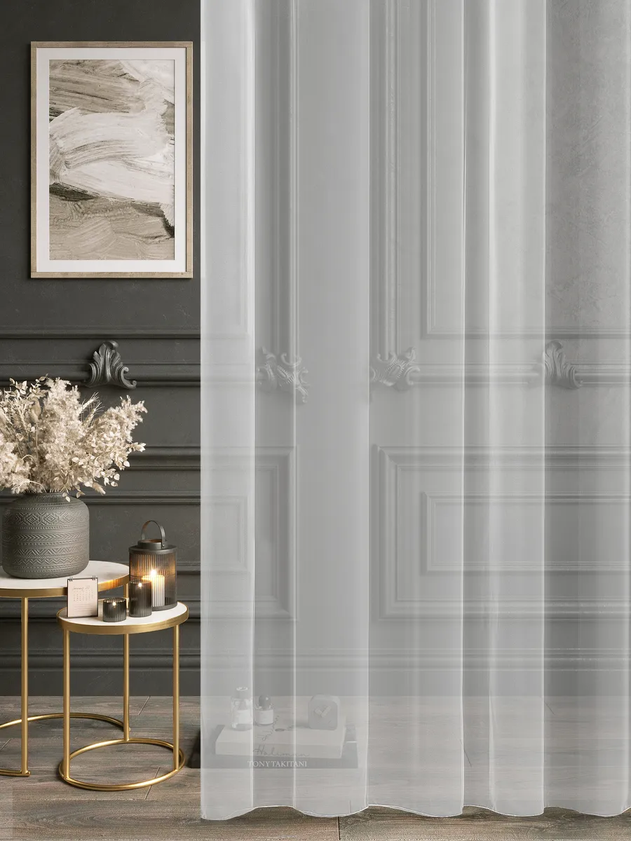 A sheer, luminous tulle curtain in soft grey tones gently drapes beside an ornate door, diffusing daylight and framing a modern side table with dried flowers and gold accents.