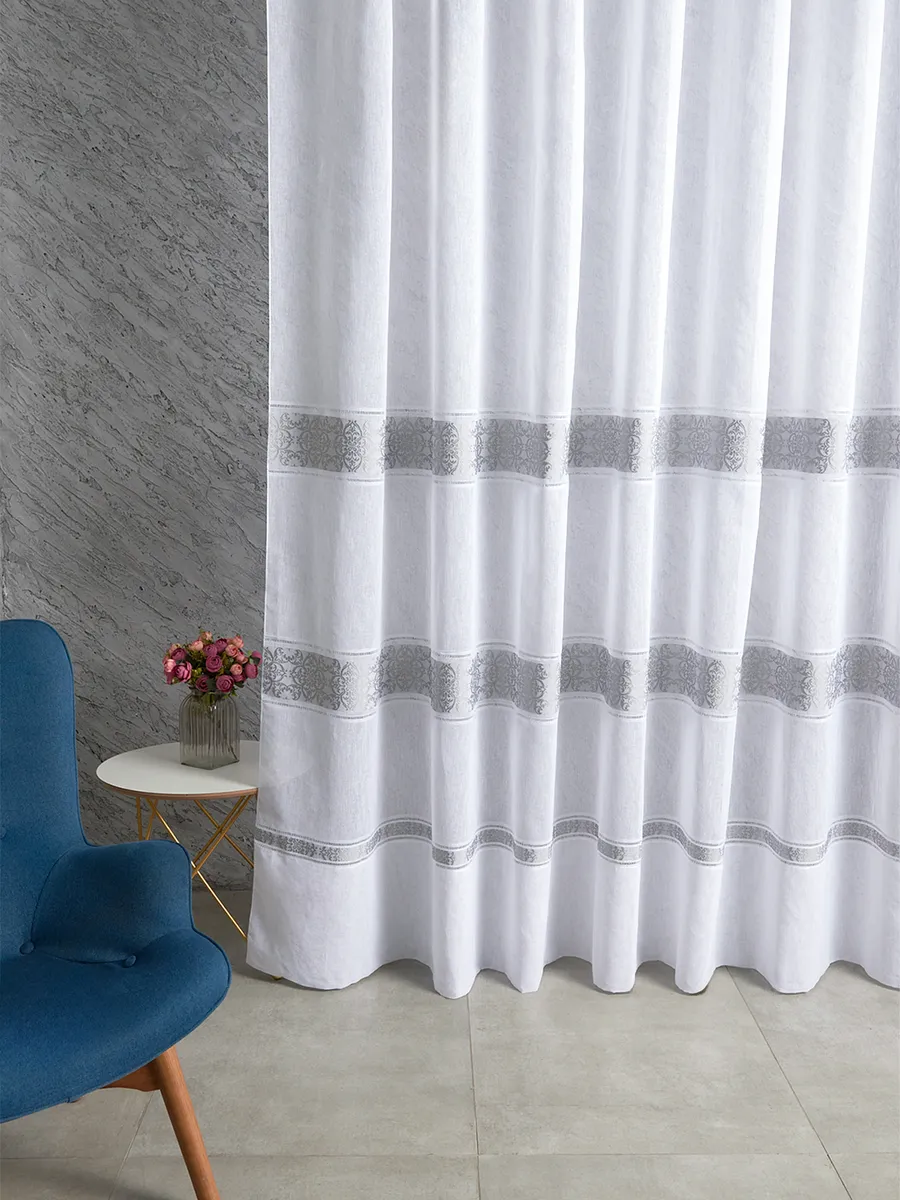 A soft white sheer curtain with silver lace bands hangs gracefully beside a blue velvet chair, diffusing natural light in a modern minimalist room with grey textured walls.