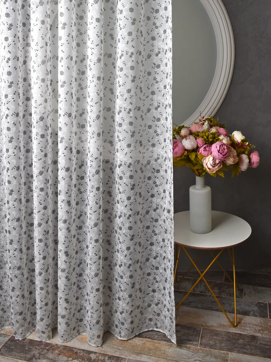 Прозрачный тюль с серыми цветочками мягко падает на пол, рядом стоит круглый столик с букетом роз в белом вазе, на фоне круглое зеркало и тёмная стена.