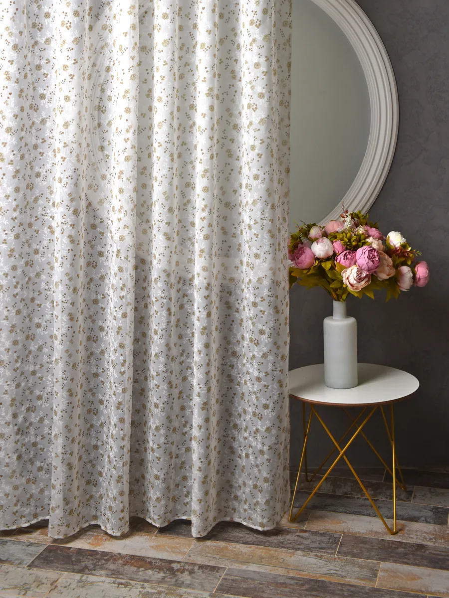 Лёгкий тюль с золотистым узором висит в светлом интерьере, рядом круглое зеркало и ваза с розовыми цветами на столике с золотыми ножками.