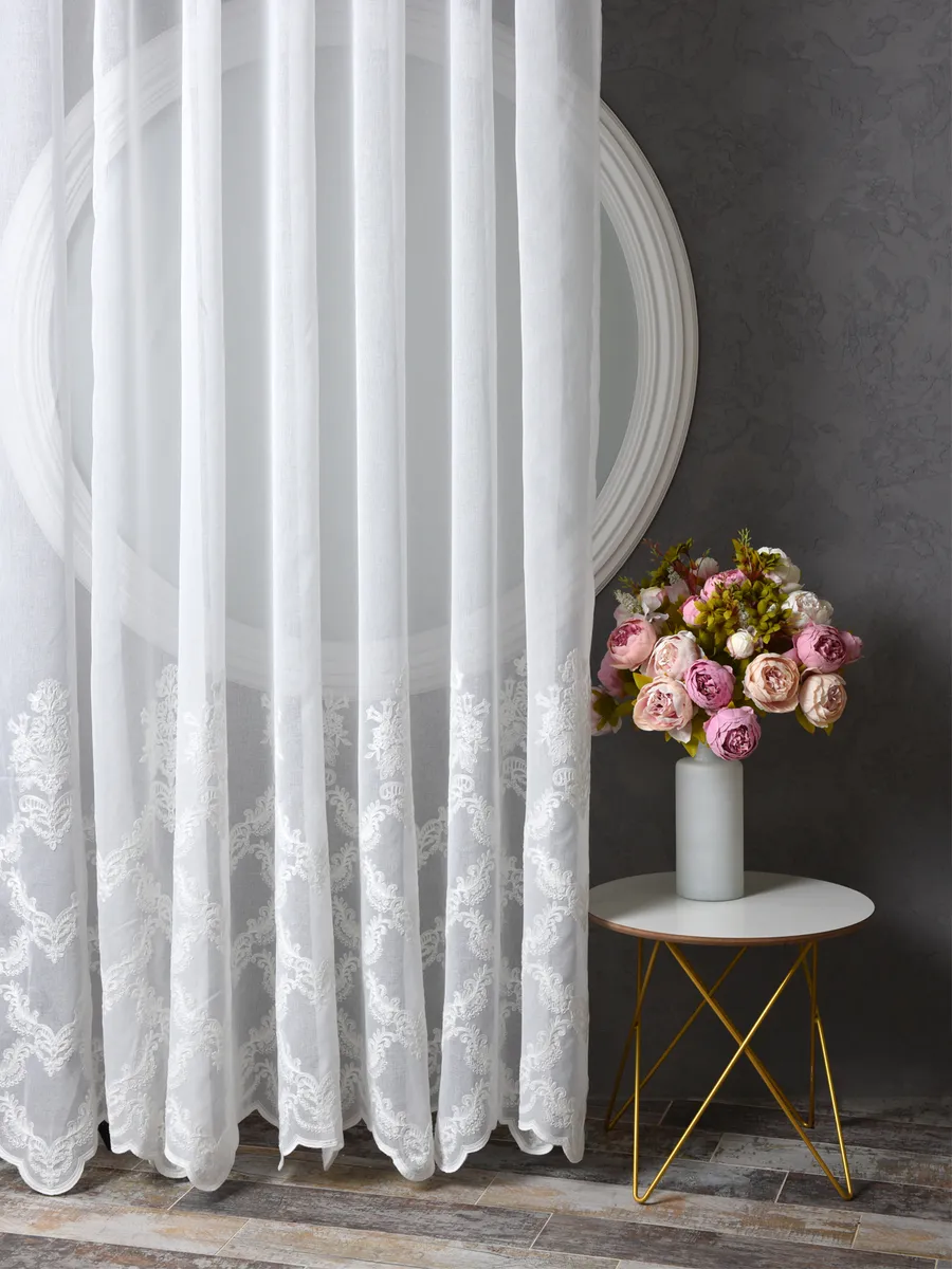 Белый тюль с кружевным узором мягко колышется у окна, за ним виден круглый зеркальный карниз, рядом — столик с букетом роз на фоне серой стены.