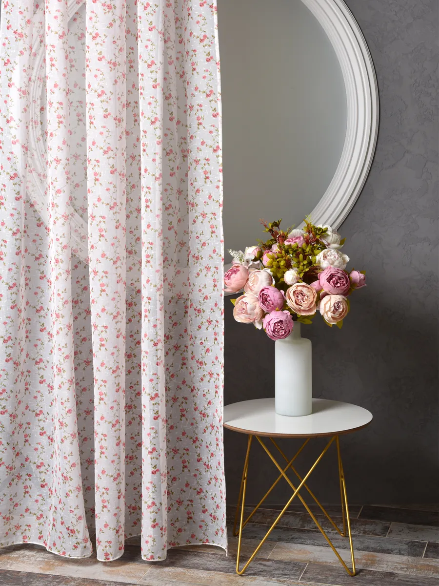 Лёгкий тюль с розовыми цветами висит у окна, рядом стоит круглый столик с вазой роз, на фоне серой стены и круглого зеркала в белой раме.