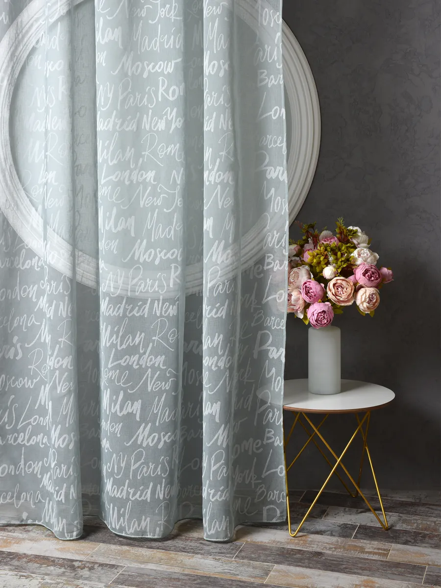 Лёгкий тюль с надписями городов висит в углу комнаты, мягко пропуская свет. Рядом — круглое зеркало и столик с букетом роз. Интерьер в серых тонах с золотыми деталями, создаёт уютную и романтичную атмосферу.