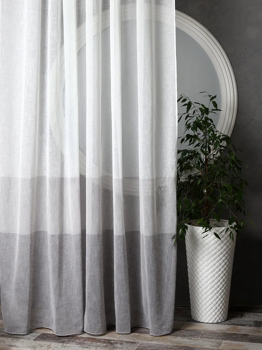 Лёгкий тюль с градиентом от белого к серому висит у окна, рядом — зелёное растение в белом вазоне, на фоне серой стены и круглого зеркала. Свет мягко проникает сквозь ткань, создавая уютную атмосферу.