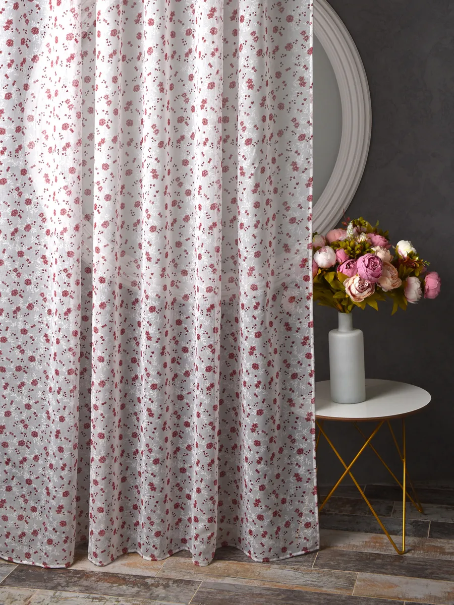 Лёгкий тюль с мелким цветочным узором висит на окне, мягко пропуская свет. Рядом — круглое зеркало и столик с букетом роз. Интерьер в тёплых тонах, уютная атмосфера, приглушенное освещение.