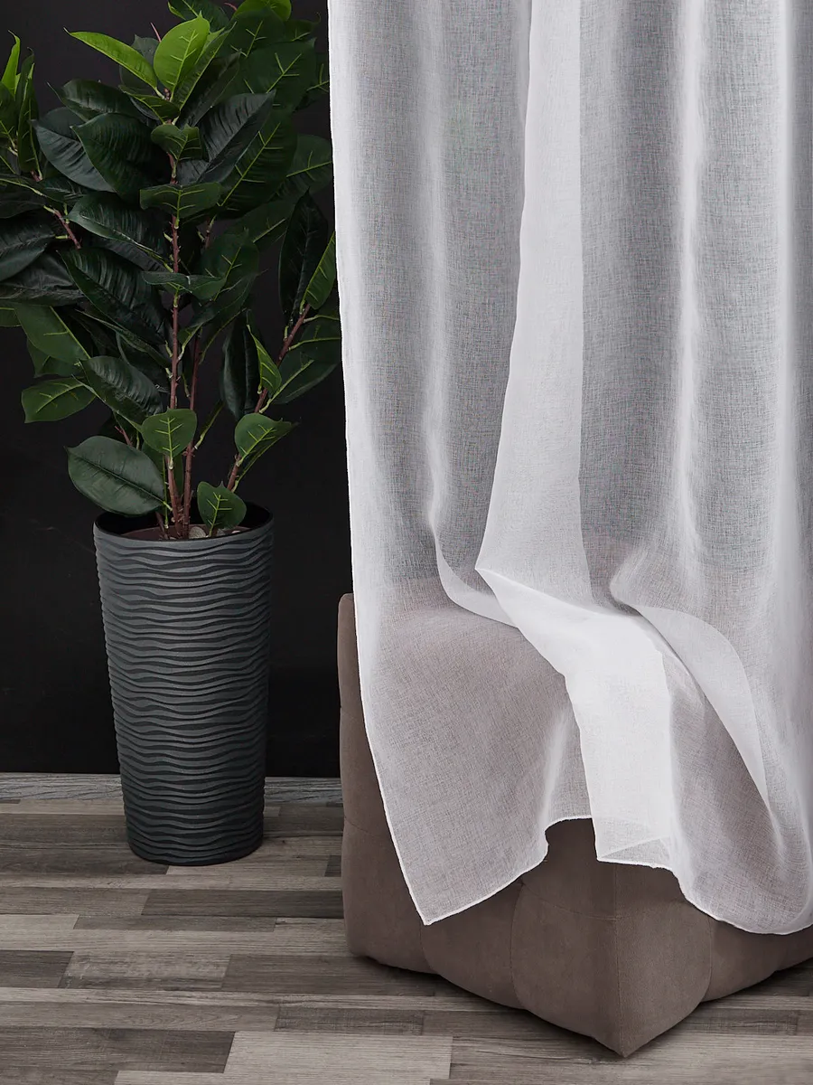 Лёгкий тюль висит на окне, мягко пропуская свет. Рядом — высокое растение в сером горшке. Интерьер в тёплых тонах, уютная атмосфера.