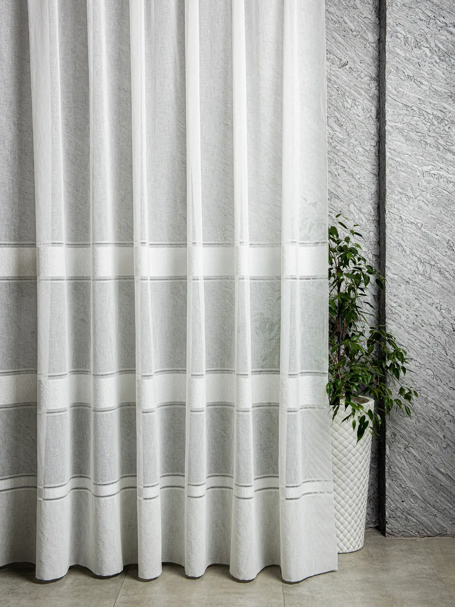 Лёгкий тюль в нежных оттенках висит на окне, мягко рассеивая свет. Рядом — зелёное растение в белом вазоне. Стена с фактурной штукатуркой. Всё в спокойных тонах, создаёт ощущение уюта и чистоты.