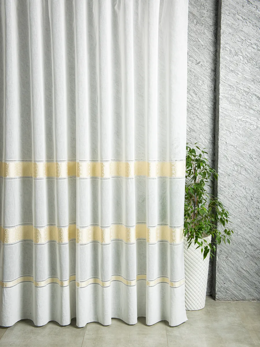 Лёгкий тюль с золотыми полосами висит на окне, мягко пропуская свет. Справа — зелёное растение в белом квадратном горшке. Стена с фактурной штукатуркой. Всё создаёт ощущение утончённой элегантности и спокойствия.