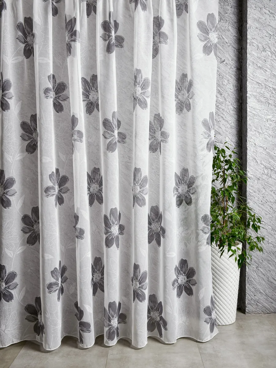 Лёгкие тюлевые шторы с серыми цветами висят на окне, рядом стоит зелёное растение в белом керамическом горшке. Интерьер в стиле минимализм с серыми стенами и светлым полом. Ткань прозрачная, рассеивает свет, создаёт уютную атмосферу.