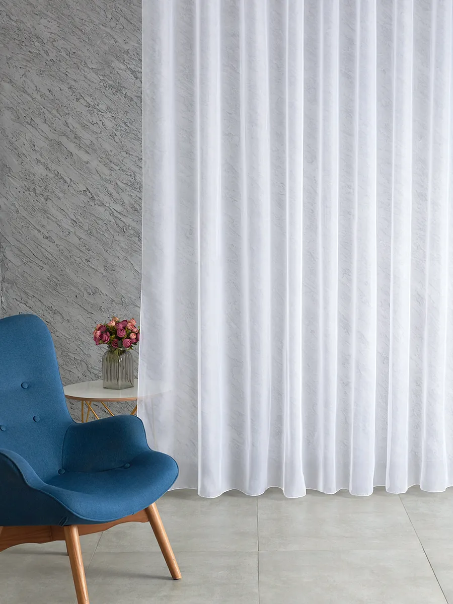 Белый тюль с мягкими складками висит на окне, пропуская рассеянный свет. Слева — синее кресло и ваза с розами на круглом столике. Стена с фактурой, пол из светлого камня. Уютная, светлая и воздушная атмосфера.