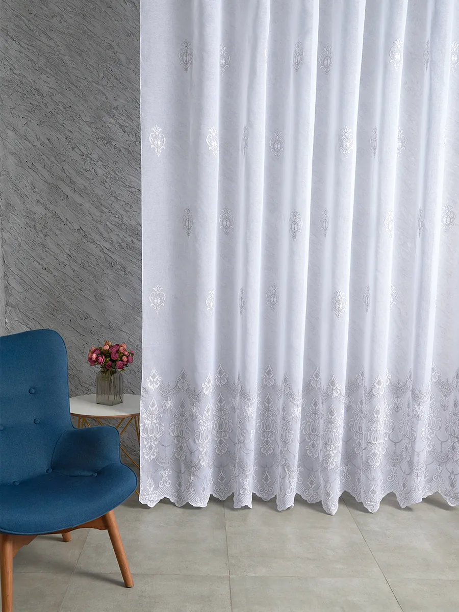 На фото — белый тюль с тонким узором и кружевным подолом, висит в светлом интерьере рядом с синим креслом и вазой с розами. Свет мягко проникает сквозь ткань, создавая атмосферу уюта и изысканности.