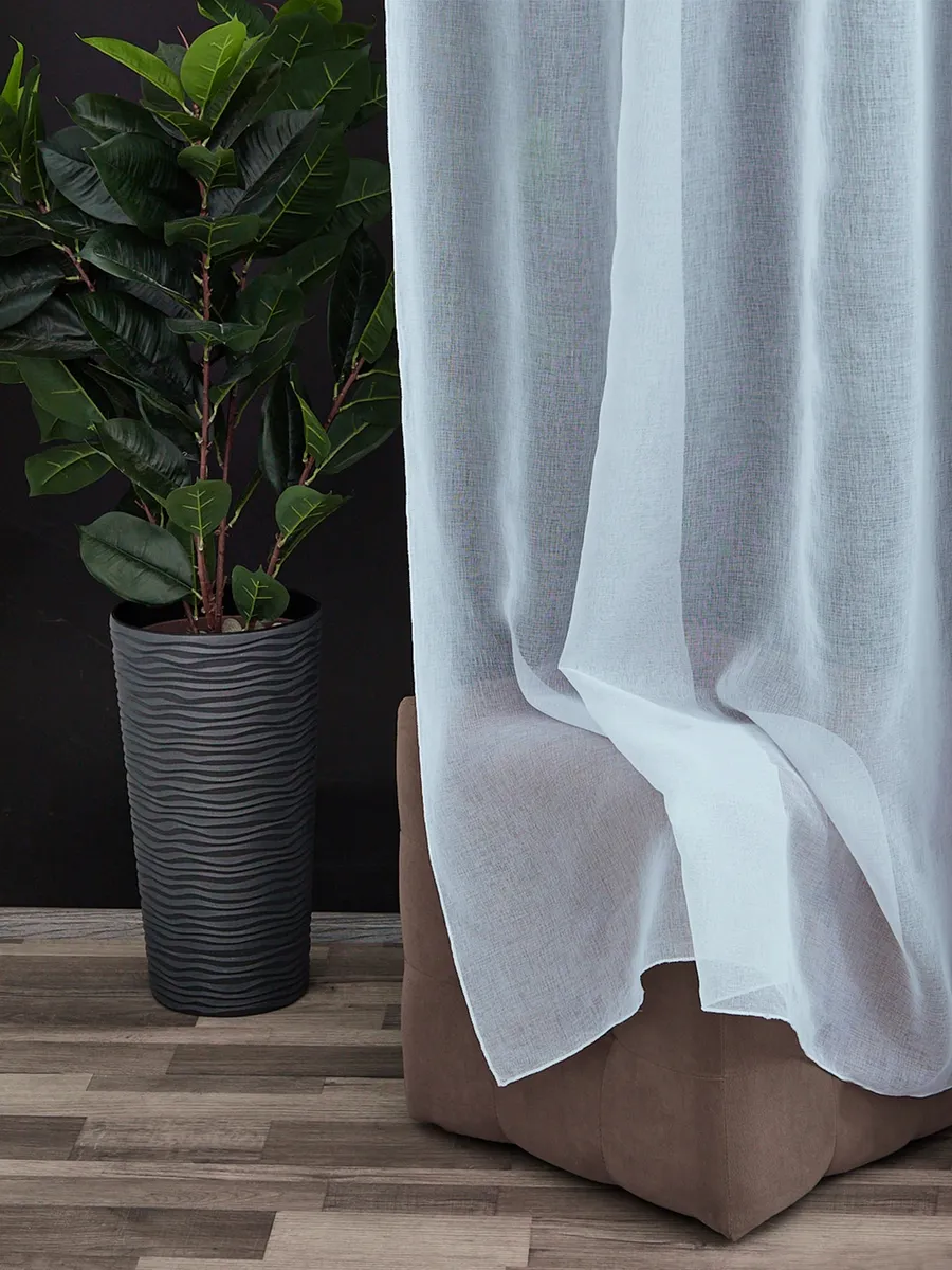 Лёгкий белый тюль плавно струится у окна, мягко фильтруя солнечный свет. Рядом — высокий тёмный горшок с зелёным растением на деревянном полу. Интерьер в тёплых тонах, спокойное утреннее освещение, атмосфера уюта и лёгкости.