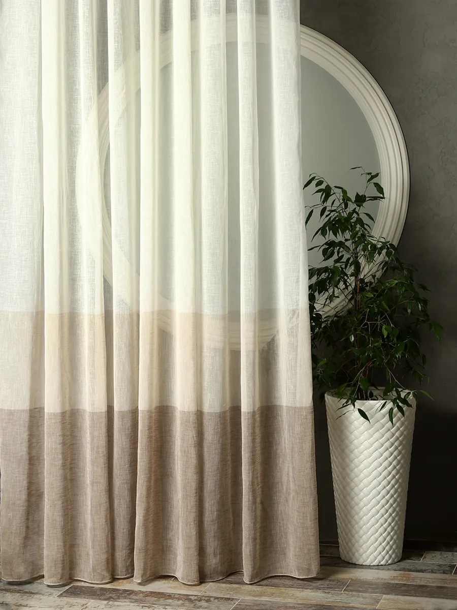 Лёгкий тюль с градиентом висит на окне, мягко пропуская свет. На заднем плане — круглое зеркало и зелёное растение в белом керамическом горшке. Интерьер в спокойных тонах, уютное освещение.