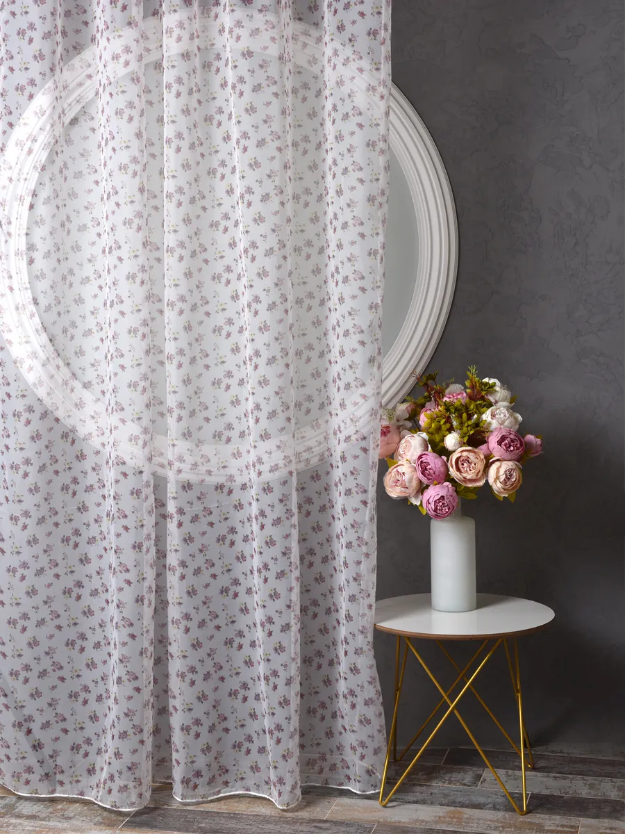 Прозрачный тюль с мелким цветочным узором висит у окна с круглым зеркалом. Розовые и белые розы в вазе на золотой ножке. Тёплый свет, серые стены, уютная атмосфера.