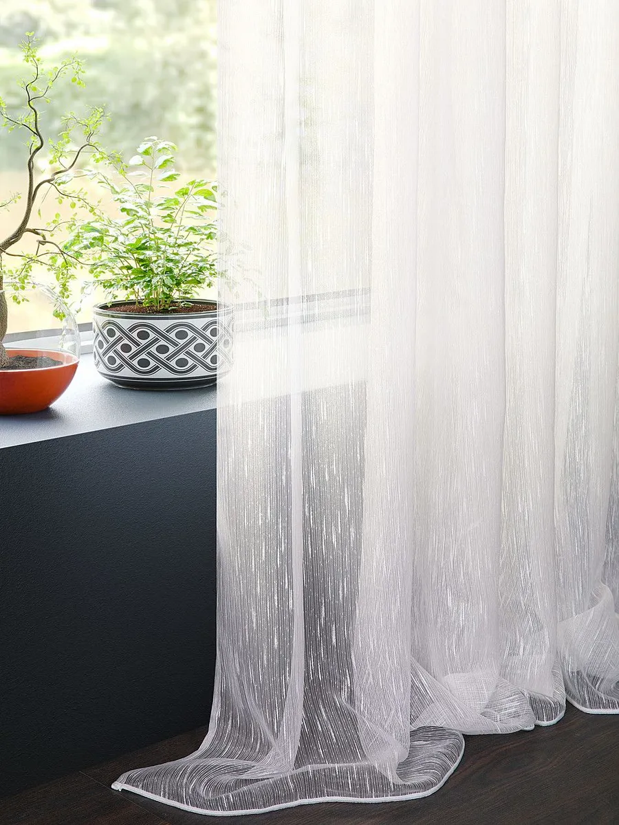 Лёгкий тюль висит на окне, мягко пропуская дневной свет. На подоконнике стоят две зелёные растения в декоративных горшках. Интерьер в тёплых тонах, настроение спокойное и умиротворяющее.