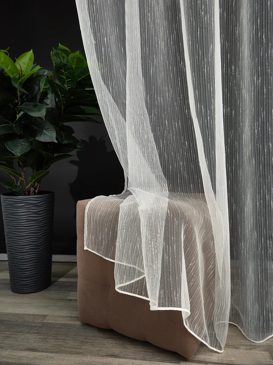 Лёгкий тюль с эффектом каскада плавно струится вдоль коричневого пуфа, рядом стоит высокий зелёный цветок в тёмном горшке на фоне чёрной стены, создаёт уютную и воздушную атмосферу.