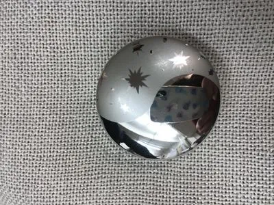 A polished metallic sphere with star cutouts rests on a textured white fabric, reflecting light and a floral pattern, evoking cosmic elegance for window decor.