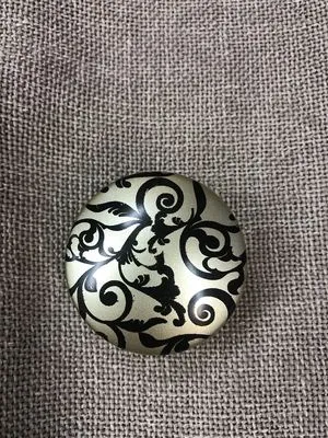 Круглый декоративный подхват с черно-серебристым узором лежит на серой льняной ткани. Виден роскошный орнамент и матовая текстура. Подходит для оконных аксессуаров в классическом стиле.