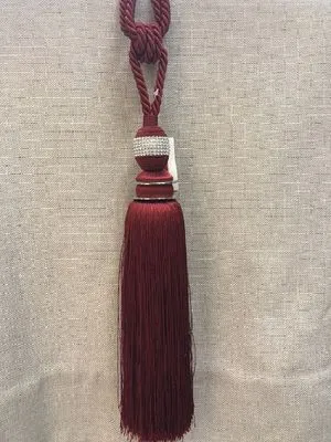Роскошный бордовый подхват с кистью и бусинами висит на фоне ткани. Декоративный элемент для штор, создающий атмосферу утончённости и роскоши в интерьере.