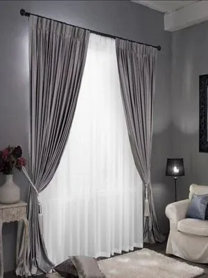 Luxurious charcoal velvet curtains with sheer white overlay, elegantly tied back in a modern gray living room with plush rug and vintage side table.