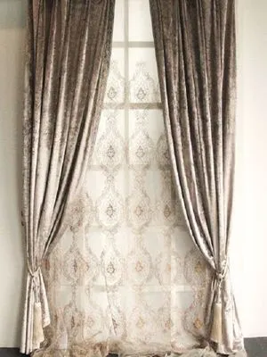 Velvet curtains in dusty taupe with gold lace overlay, elegantly tied back to reveal patterned sheers, soft natural light filtering through in a classic living room setting.