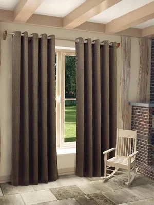 Тяжёлые бархатные шторы в тёмно-коричневом оттенке висят на люверсах, мягко скрывают окно с видом на зелёный сад. Интерьер в стиле лофт с деревянными балками, кирпичной печью и белой кресло-качалкой. Теплое естественное освещение, уютная атмосфера.