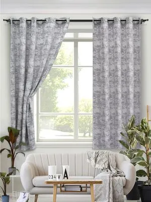 Серые шторы на люверсах в светлой гостиной, мягко падают по обе стороны от окна, за окном зелень деревьев, уютный диван с пледом и зелёные растения по бокам.