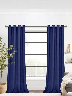 Синие бархатные шторы на люверсах висят на окне с чёрной рамой. Свет мягко фильтруется, создавая расслабляющую атмосферу. В комнате — зелёное растение в плетёном горшке и диван с белым пледом.