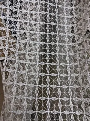 Белая сетчатая штора с геометрическим узором висит на окне, пропуская рассеянный свет. Интерьер в стиле минимализм, нейтральные тона, уютное освещение. Ткань колышется слабым ветерком, создавая игру света и тени.